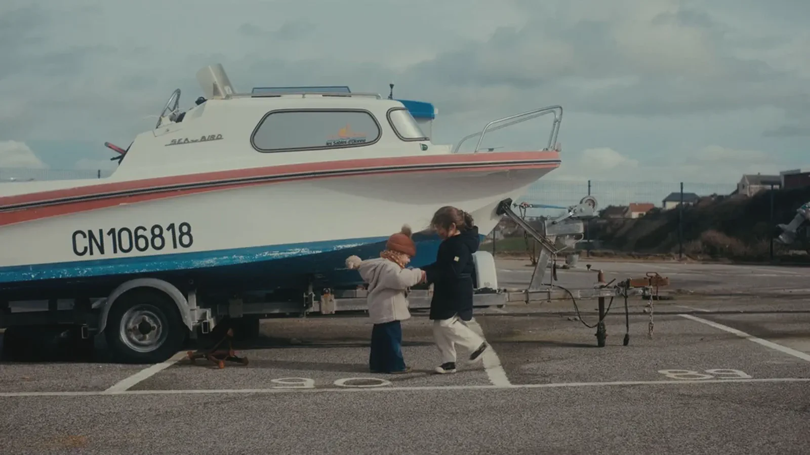 Deux enfants marchent main dans la main devant un bateau sur remorque, dans un parking au bord de mer.