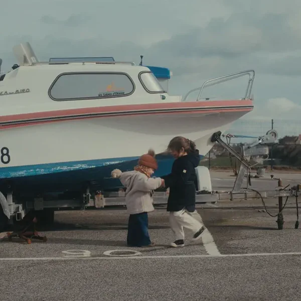Deux enfants marchent main dans la main devant un bateau sur remorque, dans un parking au bord de mer.