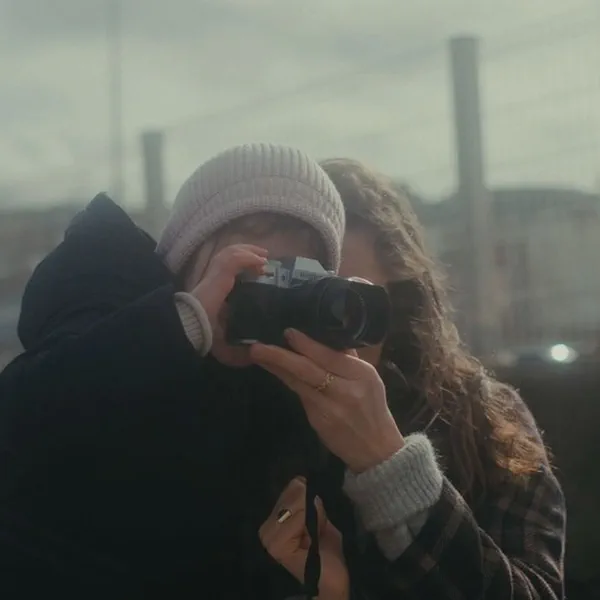 Une femme guide les mains d’un enfant coiffé d’un bonnet pour cadrer une photo, quai industriel flou en arrière-plan.