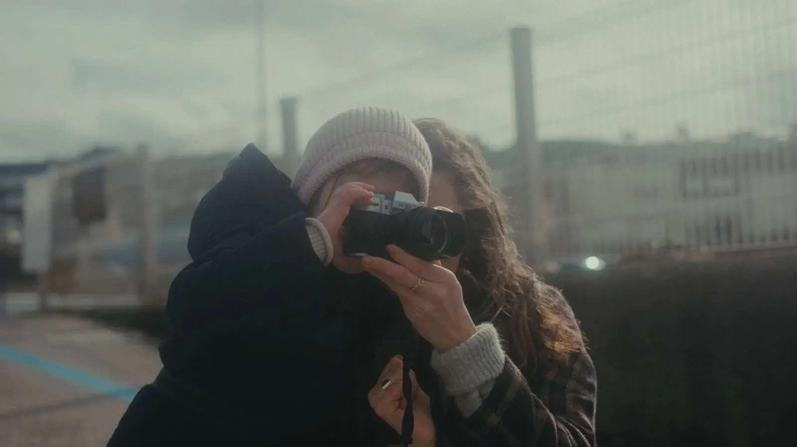Une femme guide les mains d’un enfant coiffé d’un bonnet pour cadrer une photo, quai industriel flou en arrière-plan.