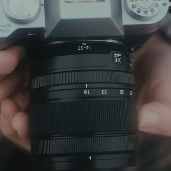 Gros plan sur un objectif zoom 16-50mm tenu à deux mains, bagues de réglage et graduations visibles.