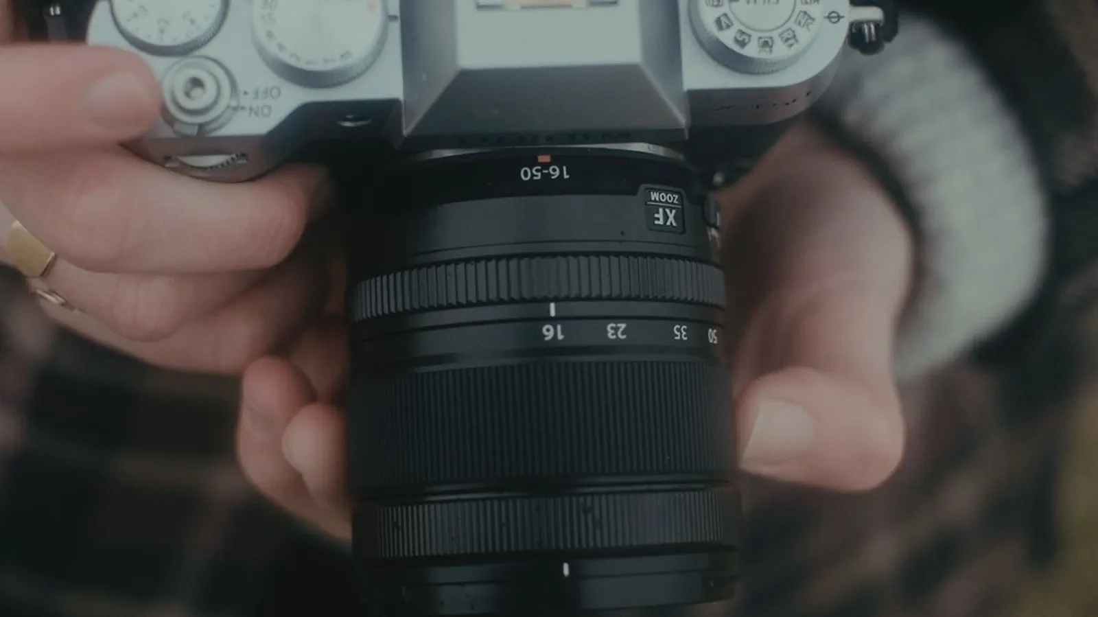 Gros plan sur un objectif zoom 16-50mm tenu à deux mains, bagues de réglage et graduations visibles.
