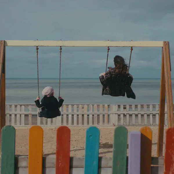 Deux personnes sur des balançoires face à la mer, vues de dos, barrière multicolore au premier plan.