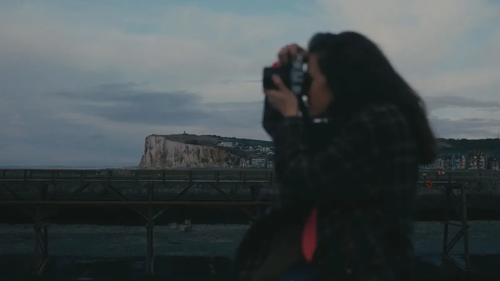 Une femme photographie des falaises blanches et une jetée au crépuscule, appareil levé devant son visage.