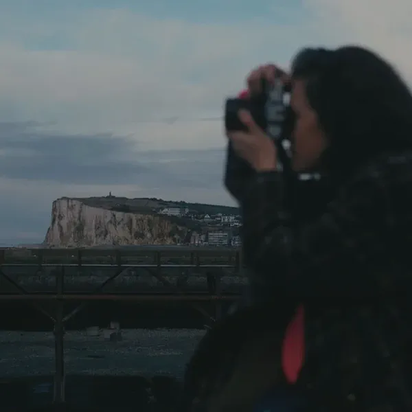 Une femme photographie des falaises blanches et une jetée au crépuscule, appareil levé devant son visage.