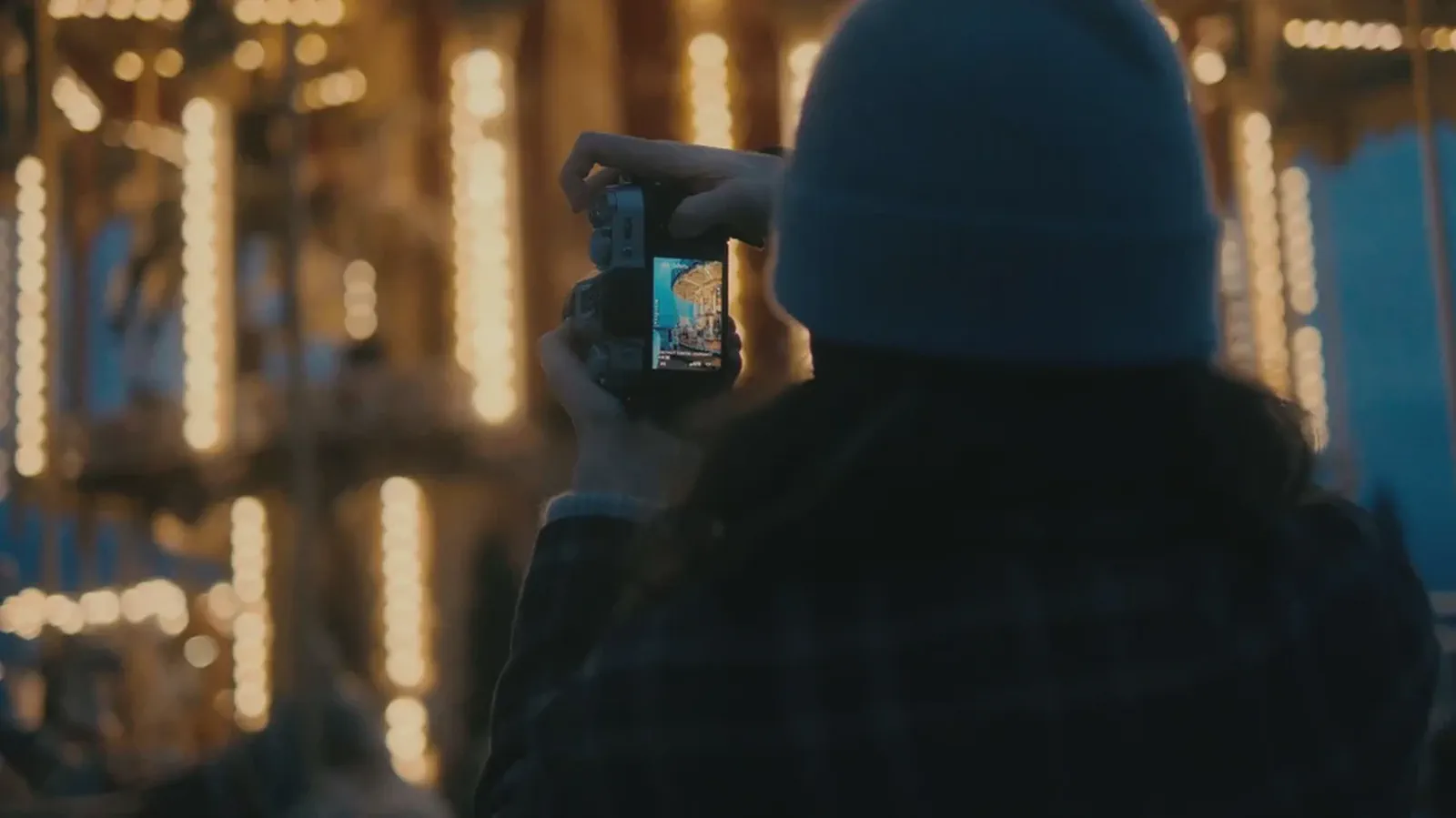Une personne de dos photographie un manège illuminé la nuit, l’écran de l’appareil affichant l’image cadrée.