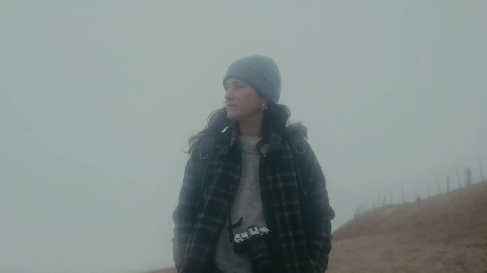 Anne Eeckeman se tient sur un chemin dans le brouillard, appareil photo autour du cou, bonnet et manteau à carreaux.