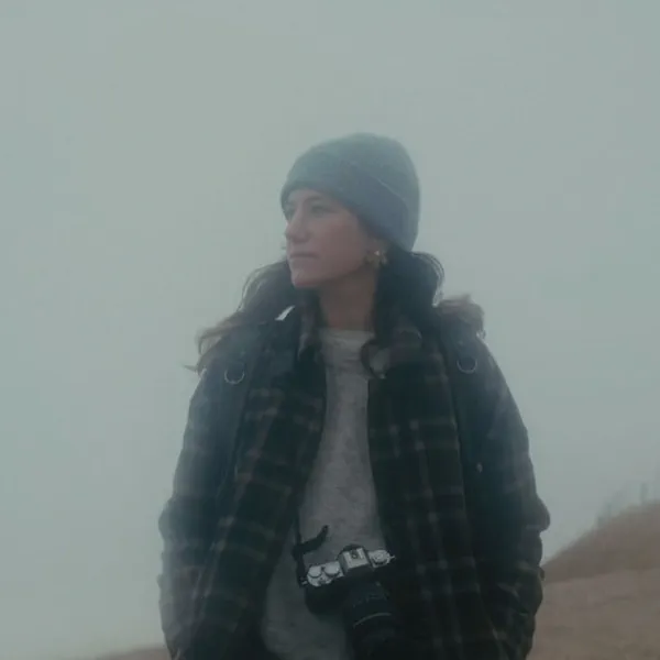 Anne Eeckeman se tient sur un chemin dans le brouillard, appareil photo autour du cou, bonnet et manteau à carreaux.