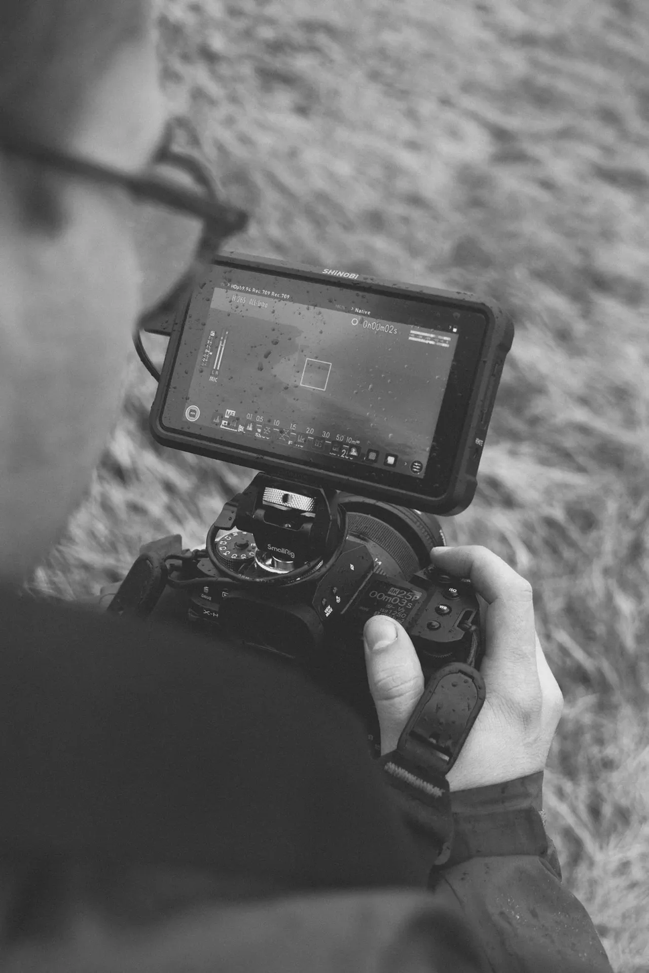 Un moniteur externe fixé sur un appareil photo affiche un cadre de visée, dunes herbeuses en arrière-plan, noir et blanc.