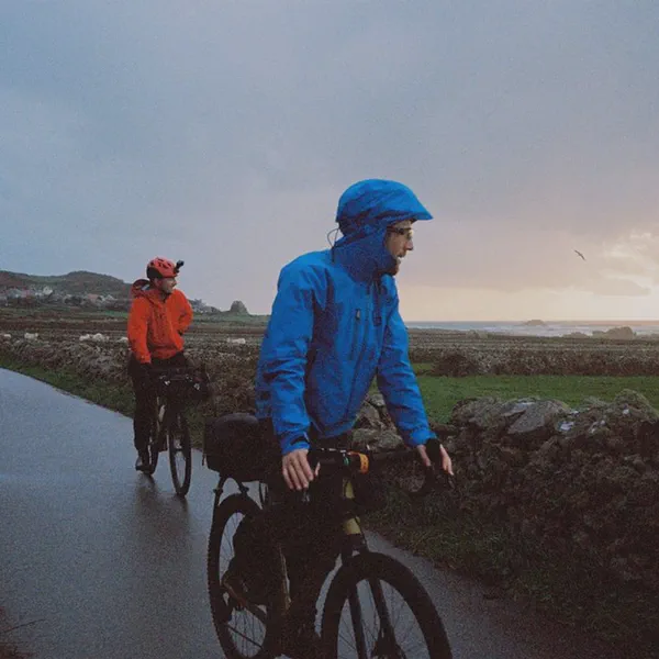 Deux cyclistes en imperméable roulent sur une route côtière au crépuscule, mer et falaises en arrière-plan.
