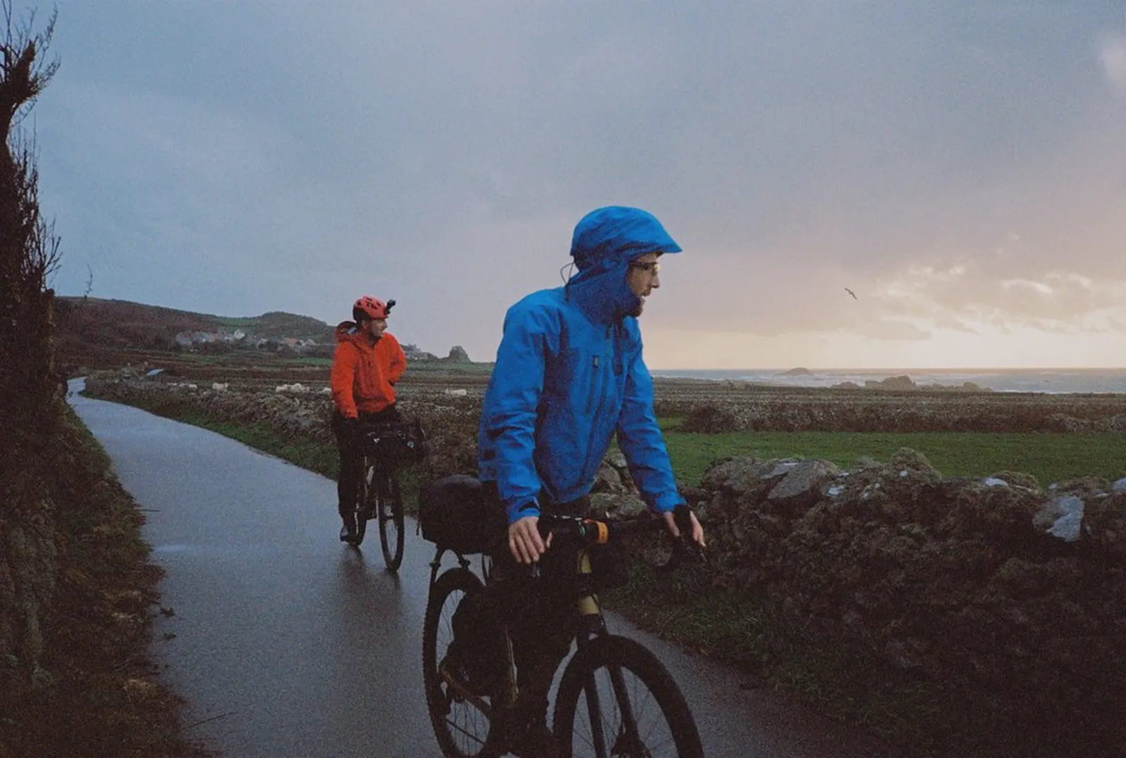 Deux cyclistes en imperméable roulent sur une route côtière au crépuscule, mer et falaises en arrière-plan.