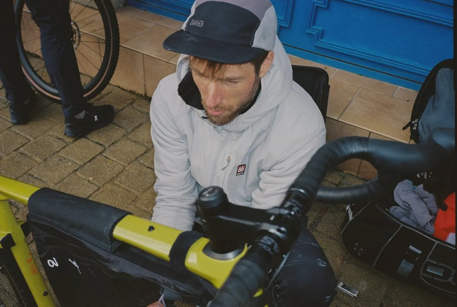 Un homme casquette règle un sac de guidon sur un vélo jaune, devant une porte bleue.