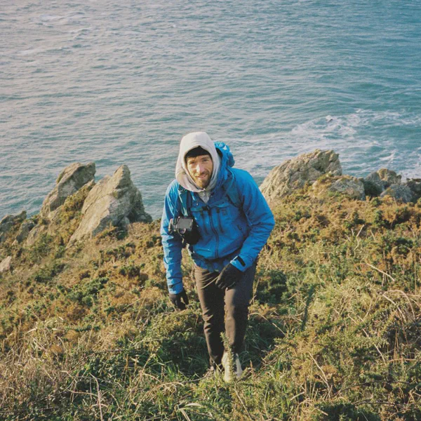 Alex Strohl gravit un sentier herbeux au bord de falaises, appareil photo autour du cou, mer en arrière-plan.