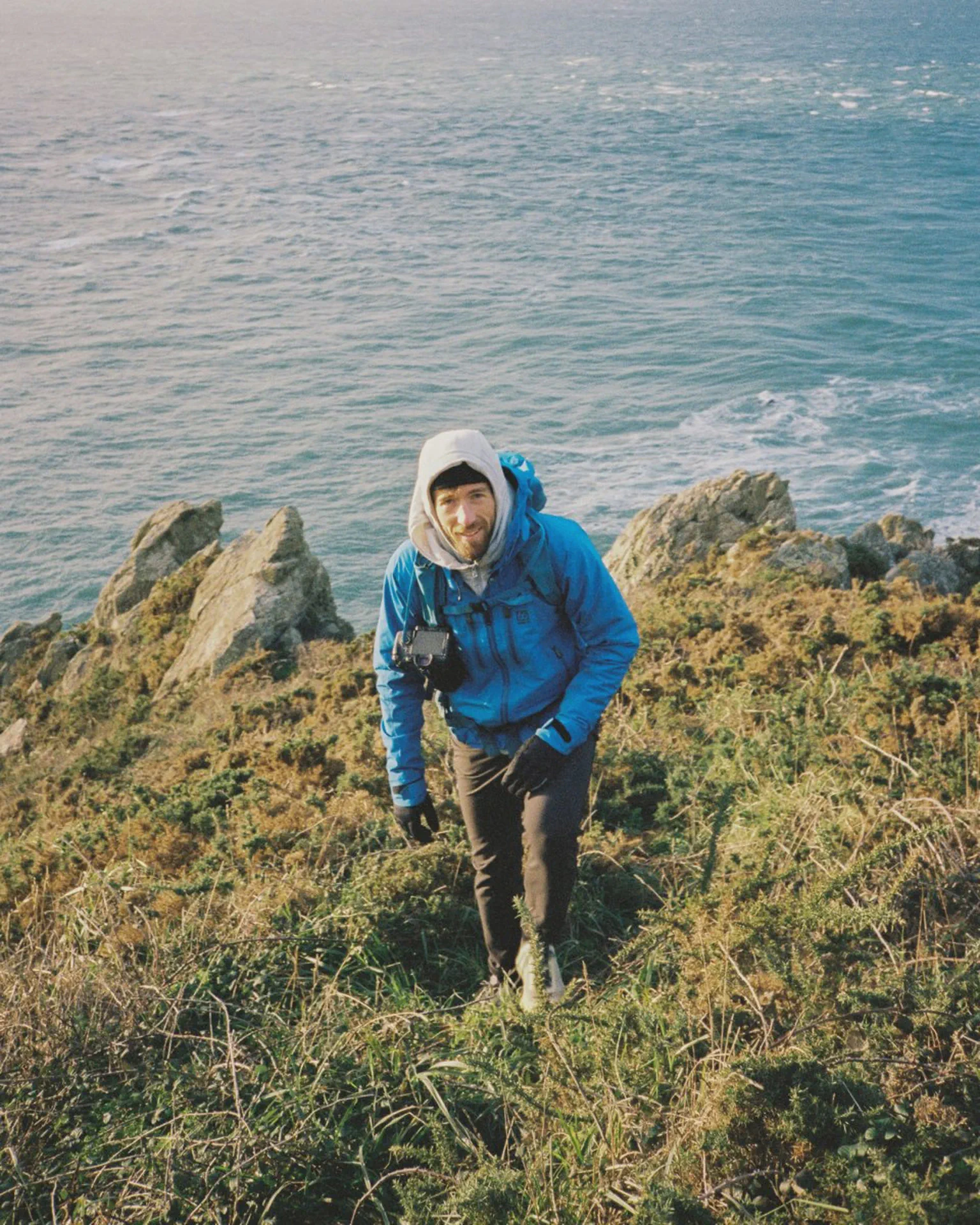 Alex Strohl gravit un sentier herbeux au bord de falaises, appareil photo autour du cou, mer en arrière-plan.