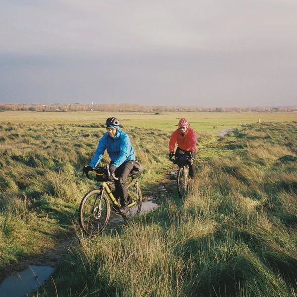 Deux cyclistes traversent un sentier herbeux marécageux, flaques d’eau au sol, village au loin.