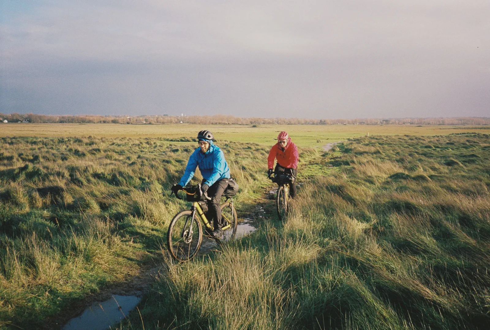 Deux cyclistes traversent un sentier herbeux marécageux, flaques d’eau au sol, village au loin.