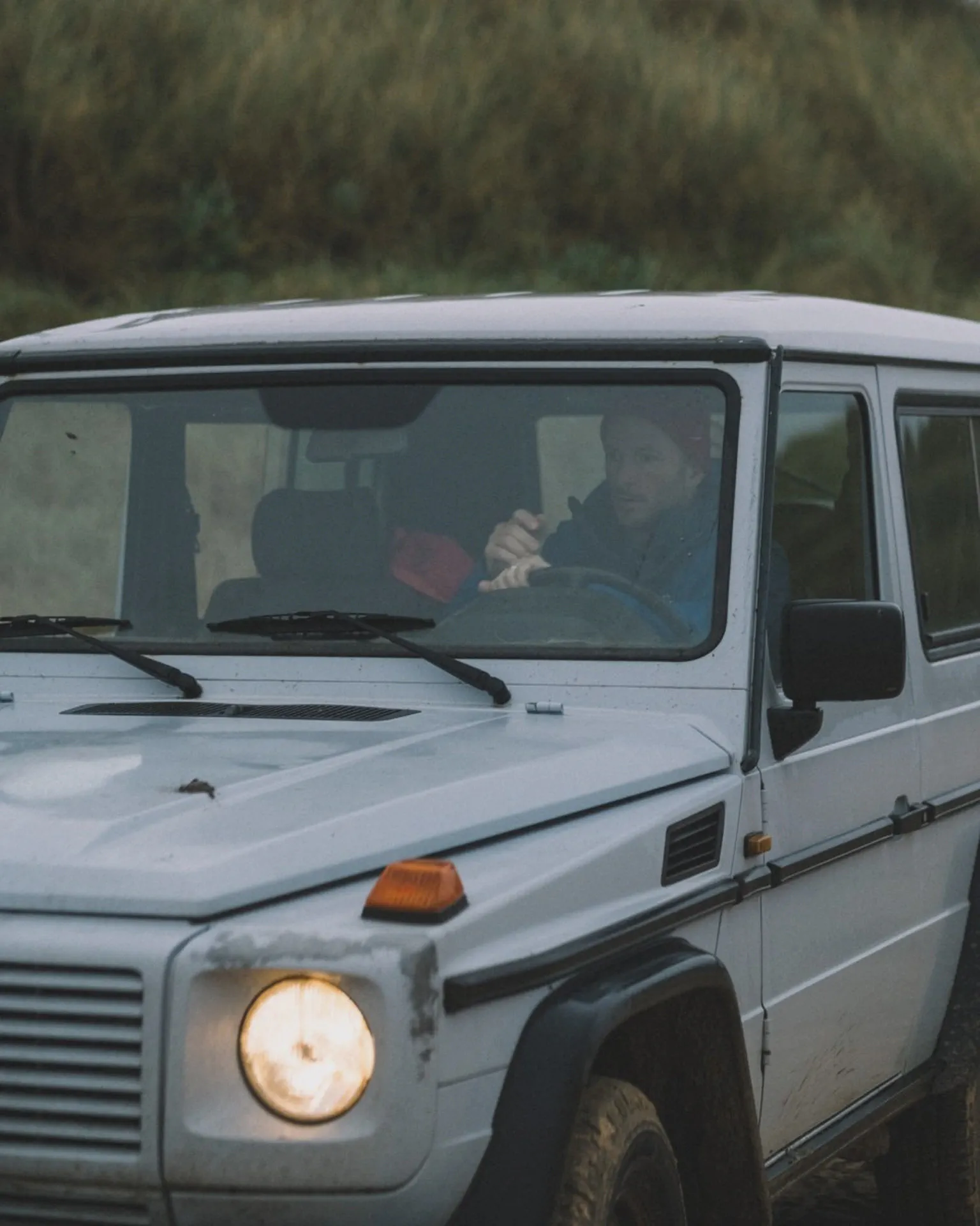 Un 4x4 blanc roule phares allumés, conducteur visible à travers le pare-brise, hautes herbes en arrière-plan.