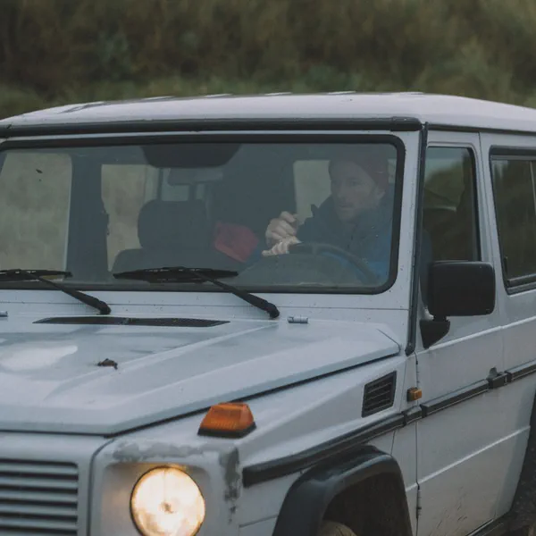 Un 4x4 blanc roule phares allumés, conducteur visible à travers le pare-brise, hautes herbes en arrière-plan.