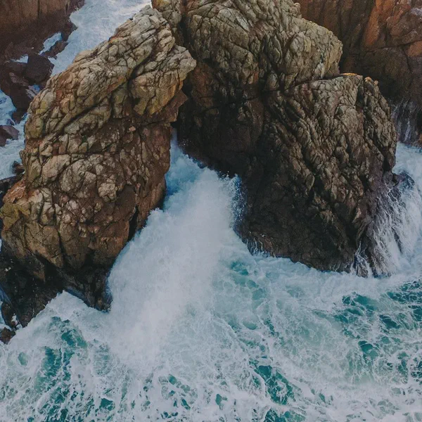 Vue aérienne de vagues qui se brisent contre des rochers escarpés, eau turquoise et écume blanche.
