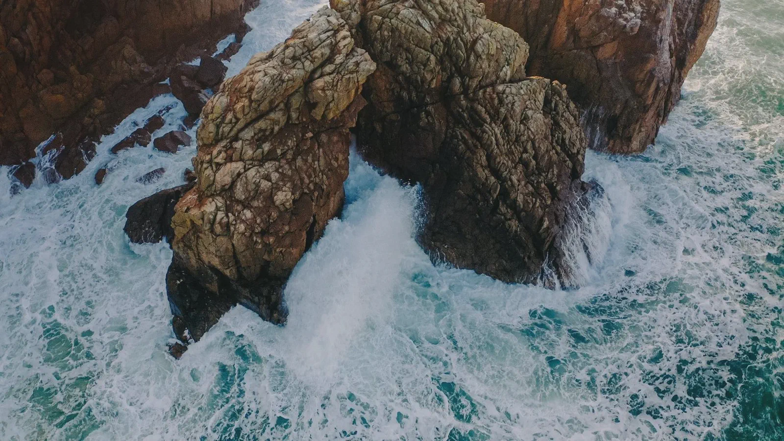 Vue aérienne de vagues qui se brisent contre des rochers escarpés, eau turquoise et écume blanche.