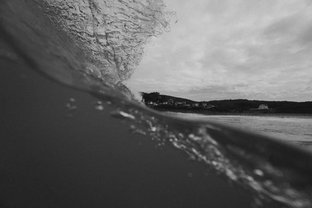 Une vague déferle en noir et blanc, embruns soulevés par le vent, côte floue à l’horizon.