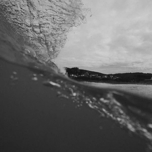Une vague déferle en noir et blanc, embruns soulevés par le vent, côte floue à l’horizon.