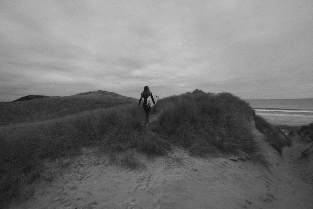 Une surfeuse marche à travers des dunes en direction de la mer, planche sous le bras, noir et blanc.
