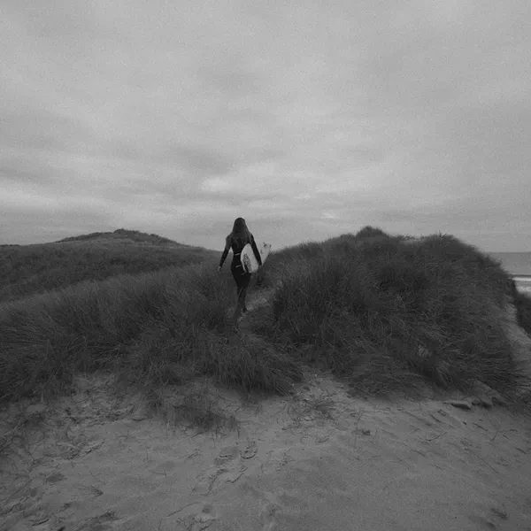 Une surfeuse marche à travers des dunes en direction de la mer, planche sous le bras, noir et blanc.