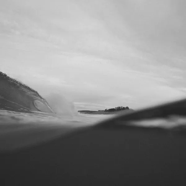 Le tube d’une vague se forme en noir et blanc, silhouette d’une pointe rocheuse visible à travers l’eau.