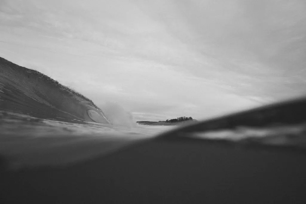 Le tube d’une vague se forme en noir et blanc, silhouette d’une pointe rocheuse visible à travers l’eau.