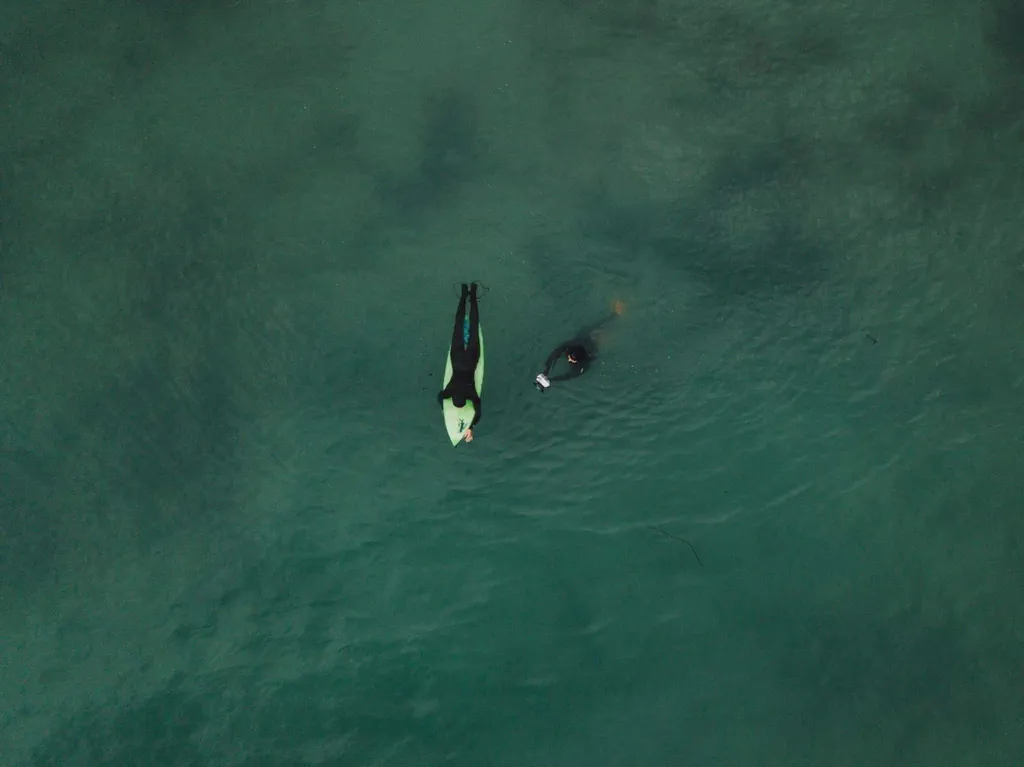 Vue aérienne de deux personnes dans une eau verte, l’une allongée sur une planche, l’autre à côté nageant.