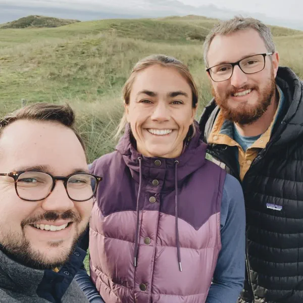 Trois personnes sourient pour un selfie devant des dunes herbeuses, ciel nuageux en arrière-plan.