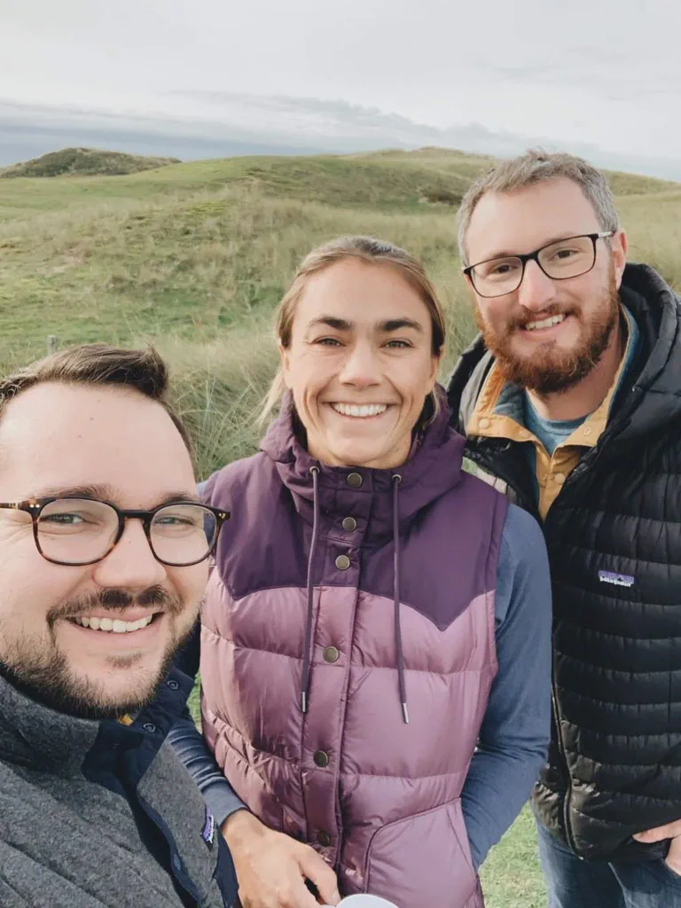 Trois personnes sourient pour un selfie devant des dunes herbeuses, ciel nuageux en arrière-plan.