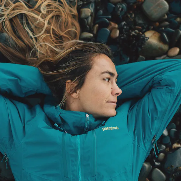 Léa Brassy allongée sur des galets, veste turquoise, cheveux étalés, regard tourné de côté.