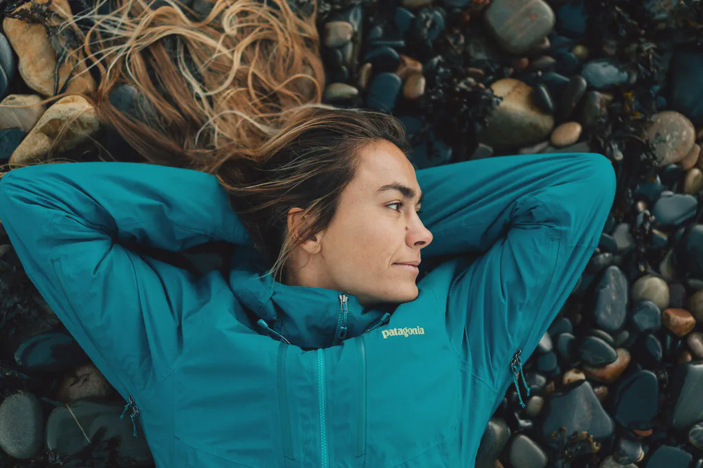 Léa Brassy allongée sur des galets, veste turquoise, cheveux étalés, regard tourné de côté.