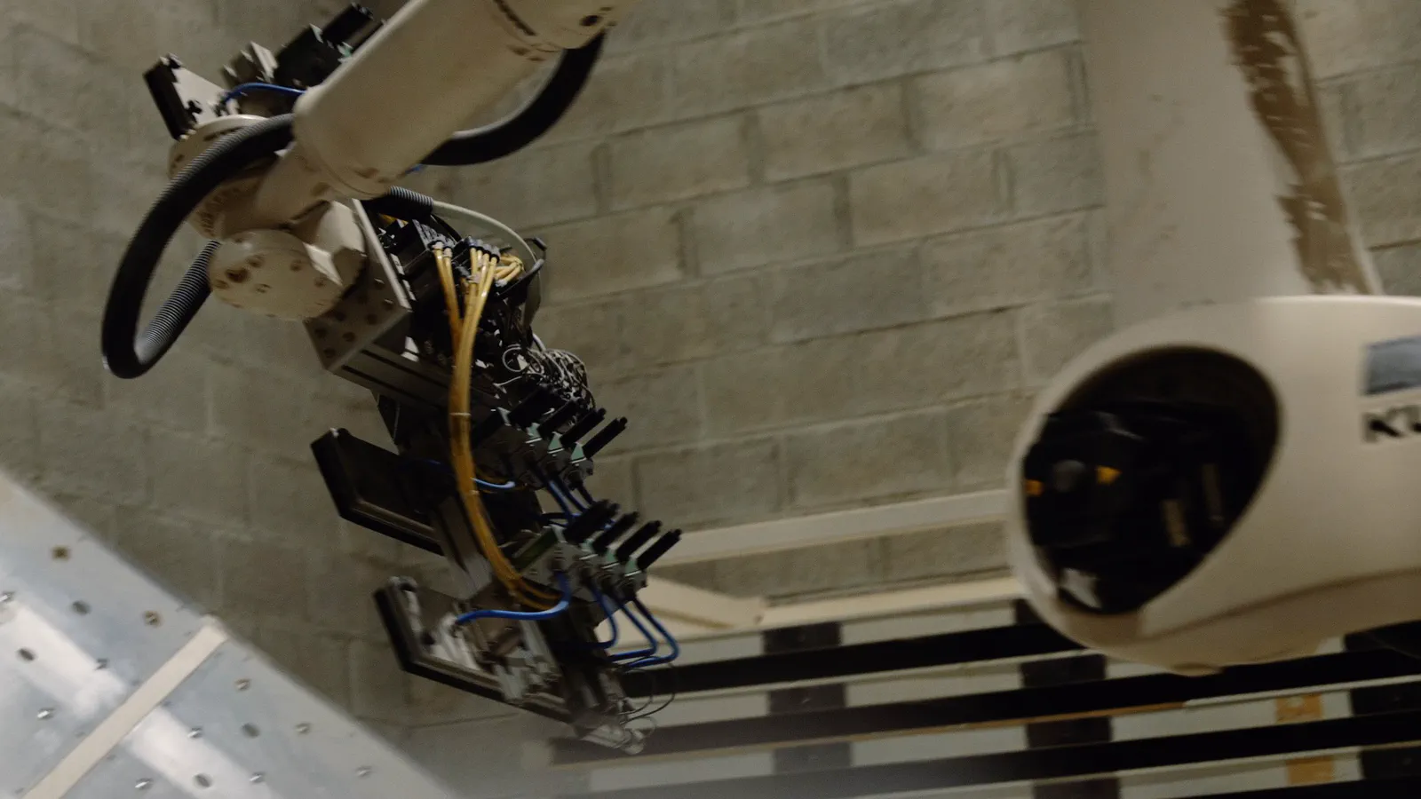Gros plan sur un bras robotisé multi-broches suspendu près d’un mur en parpaings, câbles bleus et jaunes visibles.