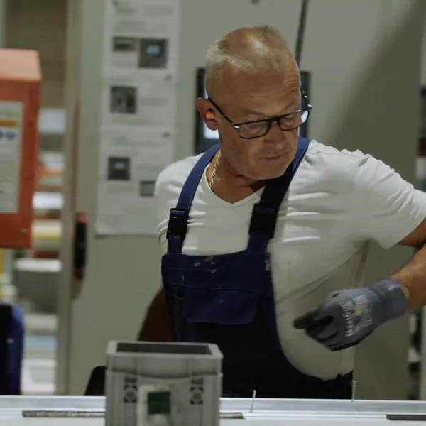 Un ouvrier chauve à lunettes en salopette bleue travaille sur une pièce métallique, gant de protection à la main.