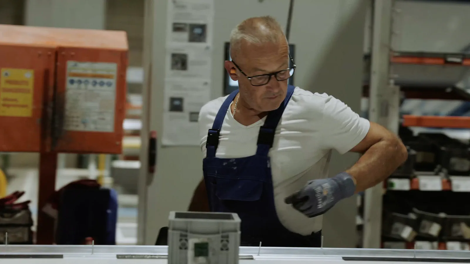 Un ouvrier chauve à lunettes en salopette bleue travaille sur une pièce métallique, gant de protection à la main.