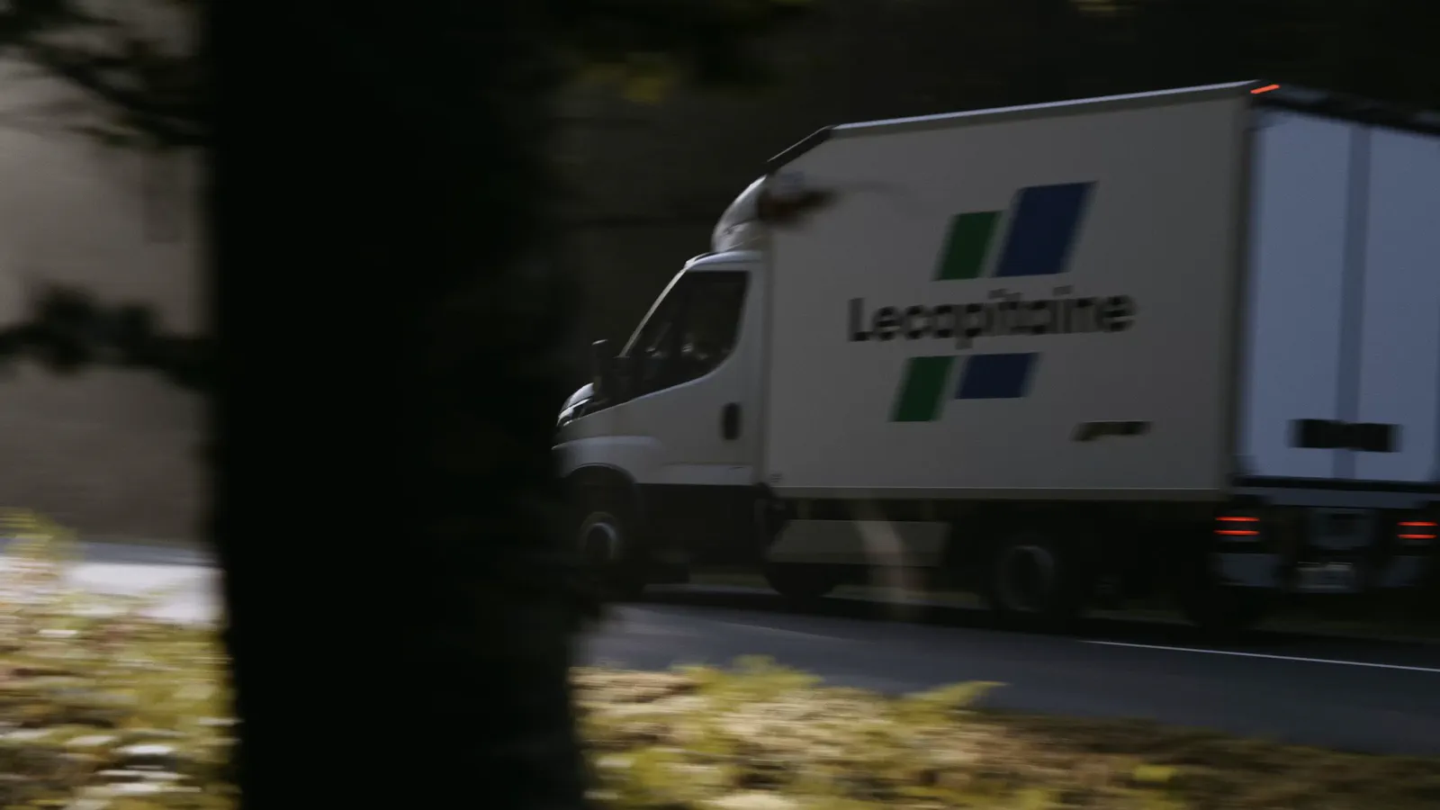 Un camion frigorifique blanc roule sur une route bordée d’arbres, vu en flou de mouvement depuis un fourré.