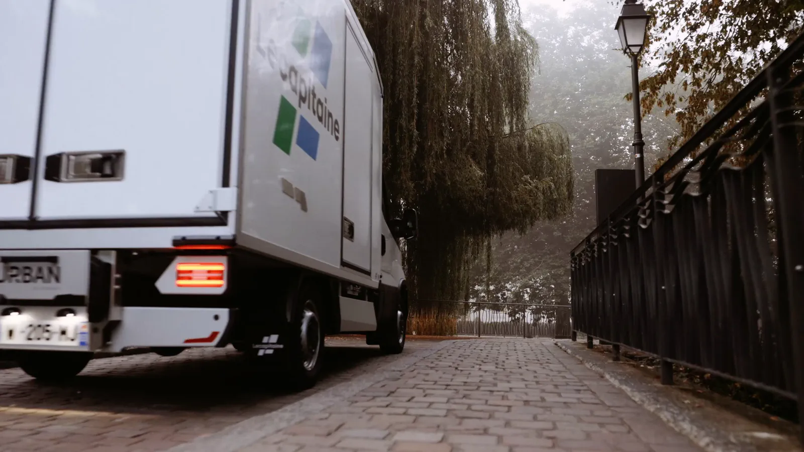 Un camion frigorifique blanc est garé sur un chemin pavé bordé d’une grille en fer forgé et d’un saule pleureur.