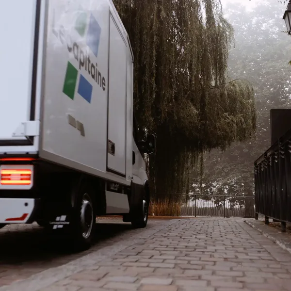 Un camion frigorifique blanc est garé sur un chemin pavé bordé d’une grille en fer forgé et d’un saule pleureur.
