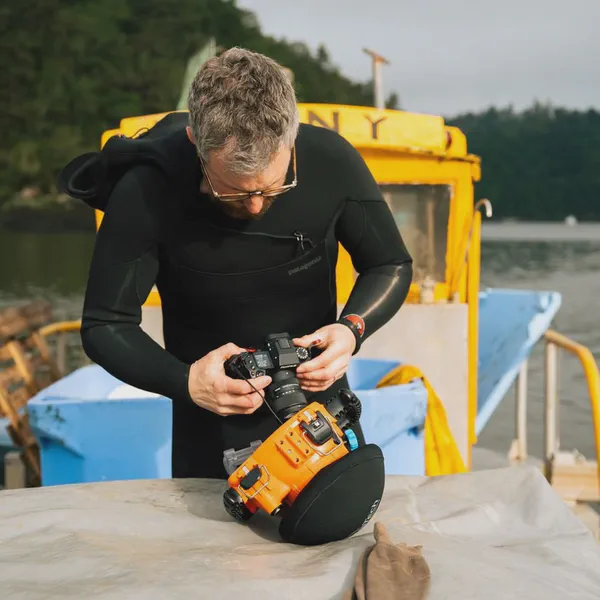 Yves Quéré règle un appareil photo fixé à un boîtier étanche orange, combinaison de plongée noire, bateau jaune en fond.
