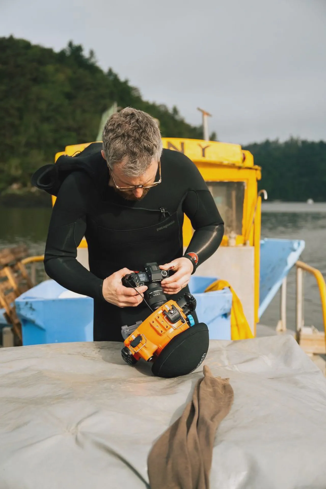 Yves Quéré règle un appareil photo fixé à un boîtier étanche orange, combinaison de plongée noire, bateau jaune en fond.