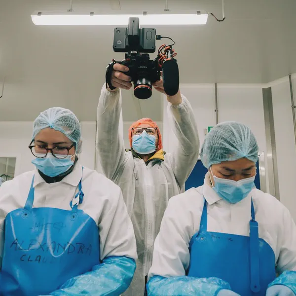 Un cadreur masqué filme au-dessus de deux employées en charlotte et masque dans un atelier agroalimentaire.