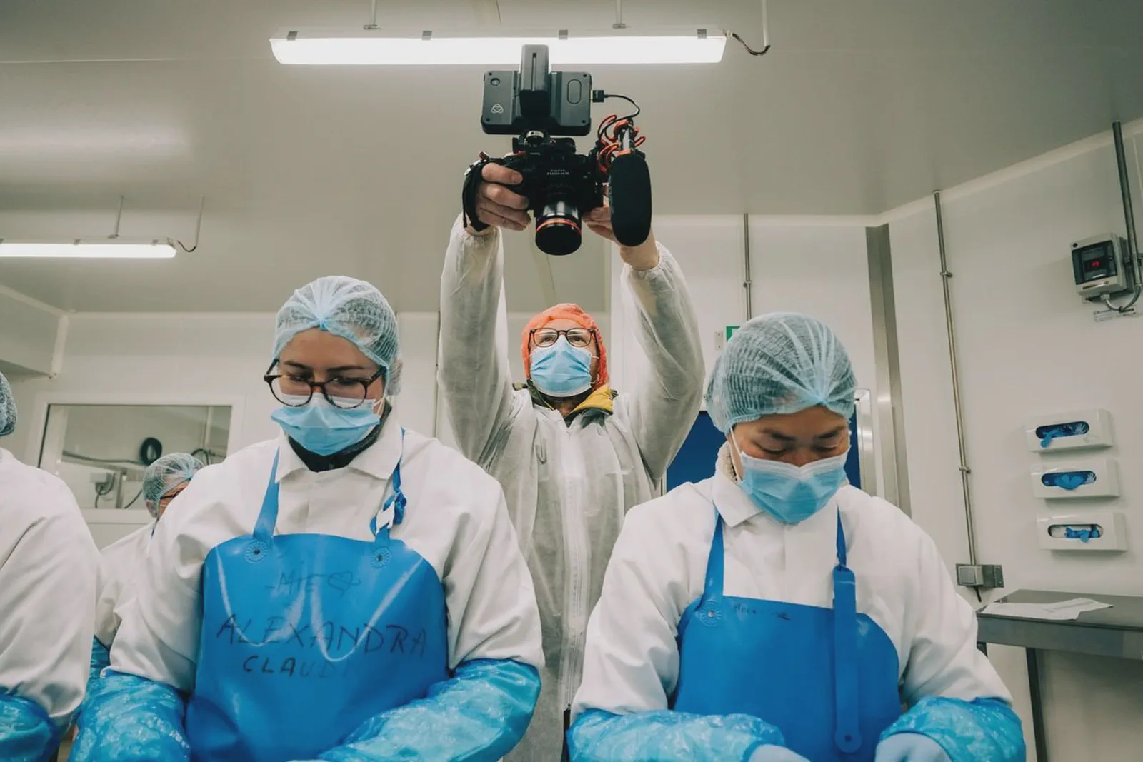 Un cadreur masqué filme au-dessus de deux employées en charlotte et masque dans un atelier agroalimentaire.