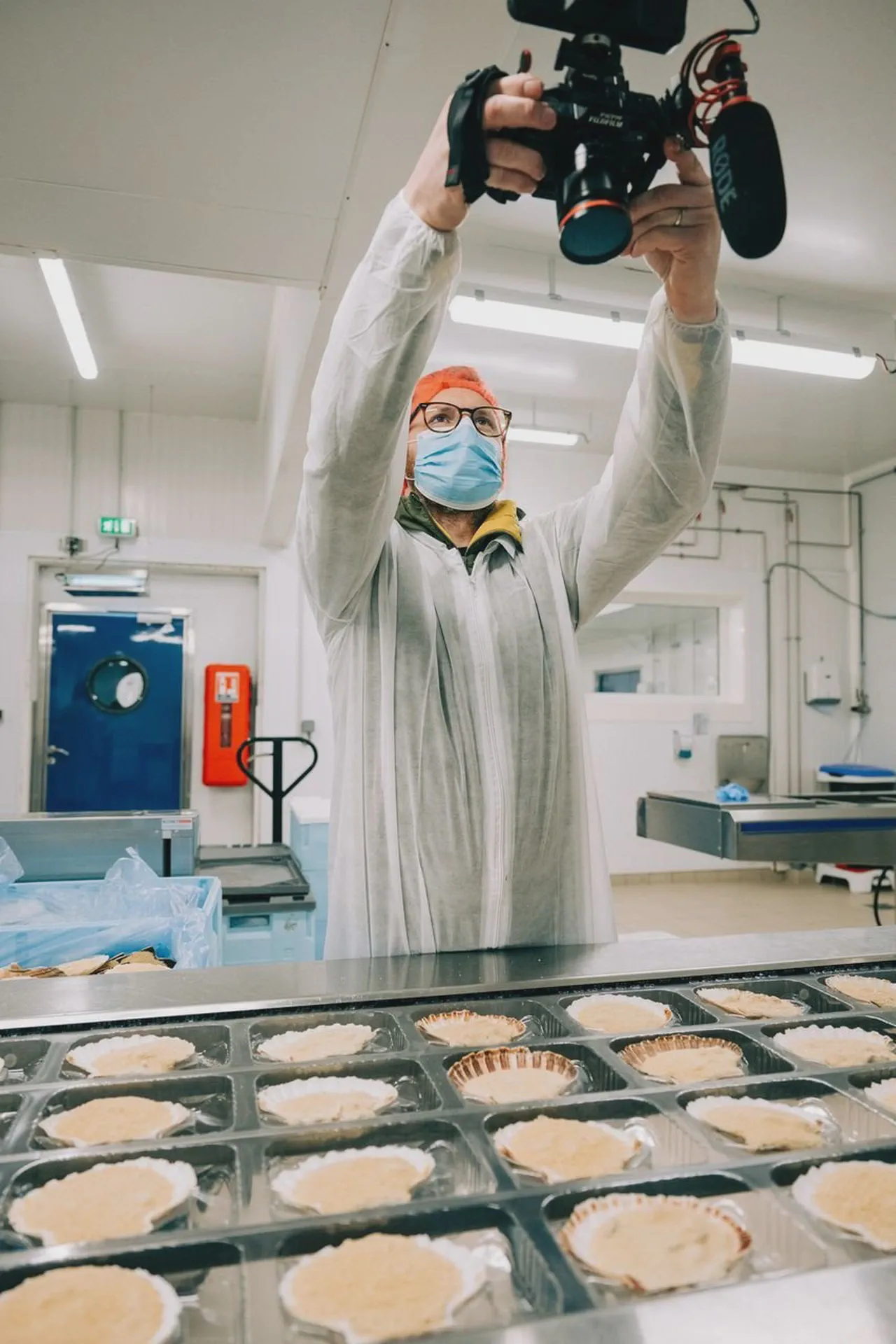 Un cadreur masqué et bonnet orange filme des noix de coquillages disposées en barquettes sur une chaîne.