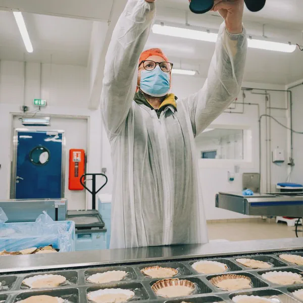 Un cadreur masqué et bonnet orange filme des noix de coquillages disposées en barquettes sur une chaîne.
