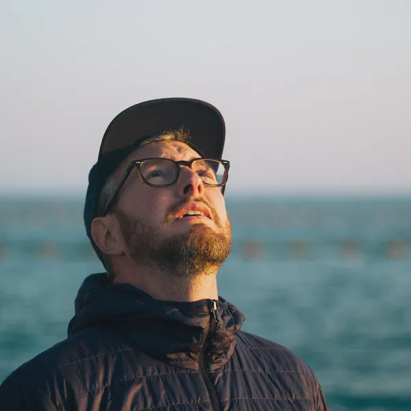 Un homme casquette et lunettes lève les yeux vers le ciel, bouées d’un parc à moules floues sur la mer derrière lui.