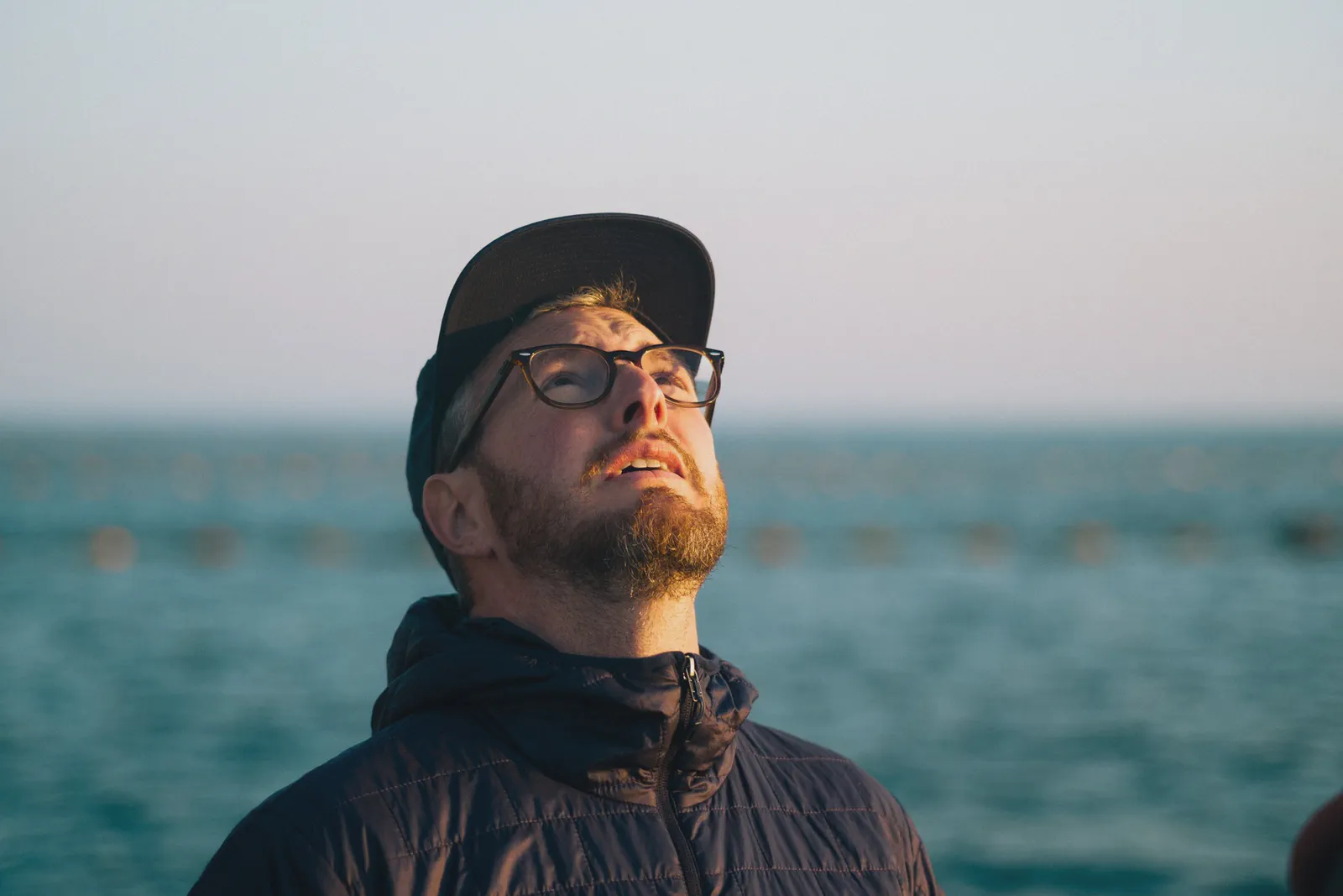 Un homme casquette et lunettes lève les yeux vers le ciel, bouées d’un parc à moules floues sur la mer derrière lui.