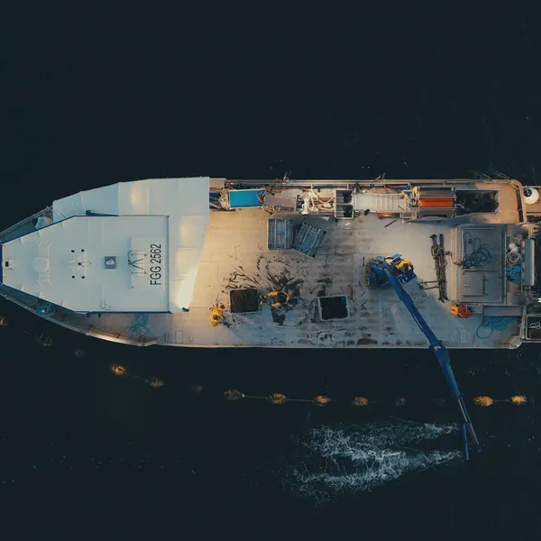 Vue aérienne du pont d’un bateau chargé de caisses et d’équipement, tuyau plongeant vers des bouées en mer.